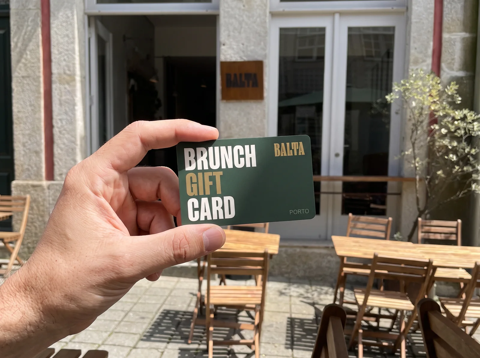 A hand holding the Balta Brunch Gift Card outside Balta, with the terrace behind